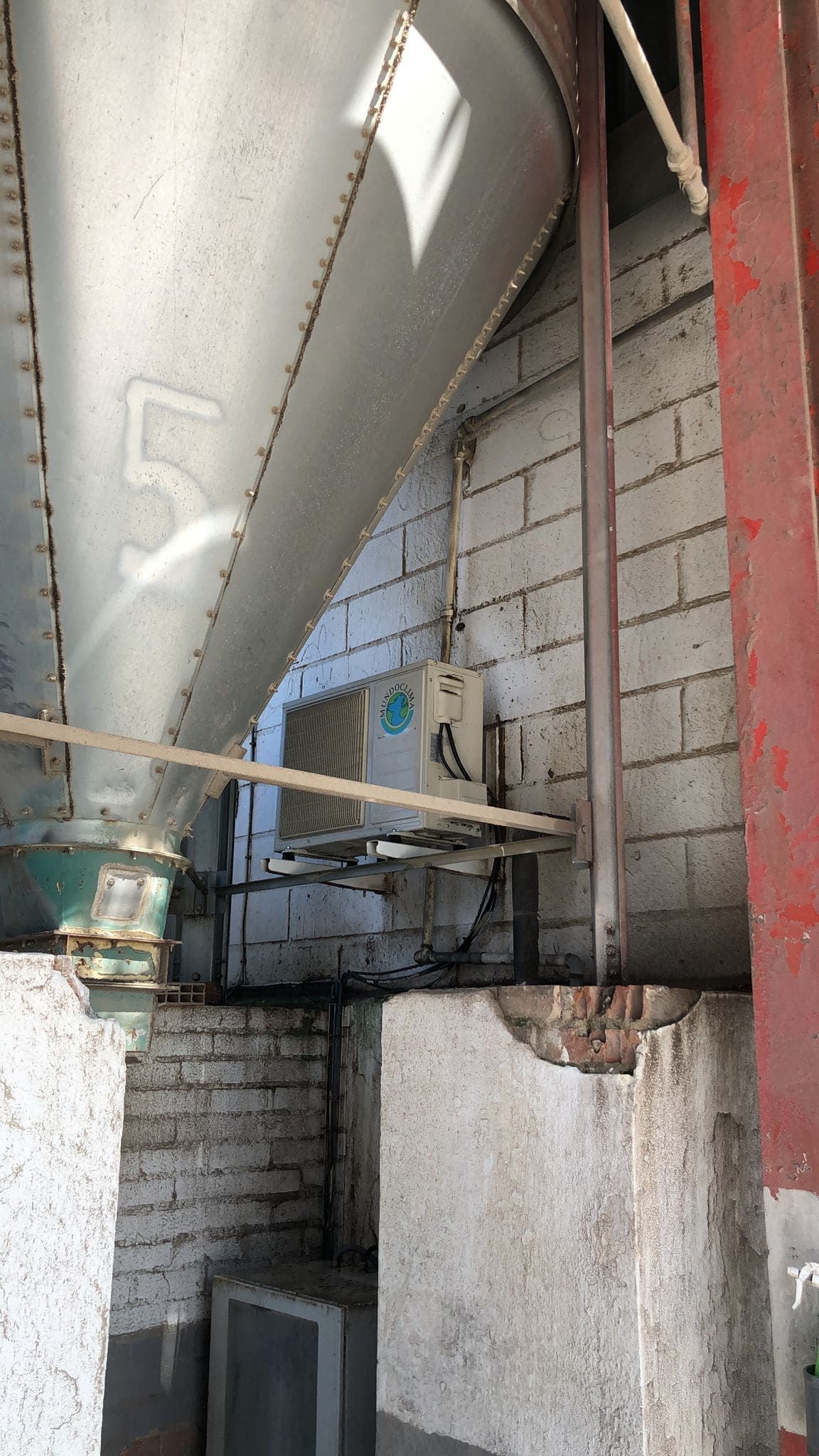 Una gran tolva industrial metálica en forma de cono, etiquetada con el número '5', se encuentra situada encima de una pared de ladrillo blanco y una unidad de aire exterior.