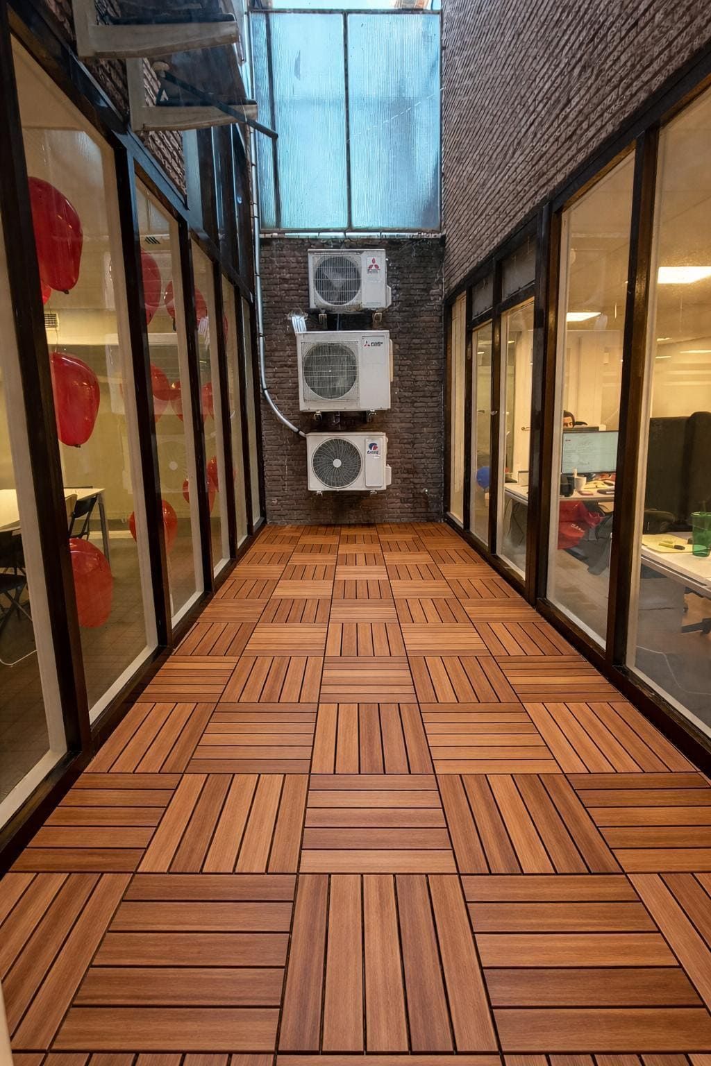 Un estrecho pasillo exterior con baldosas de madera, flanqueado por paredes de cristal que van desde el suelo hasta el techo y tres unidades de aire acondicionado apiladas.