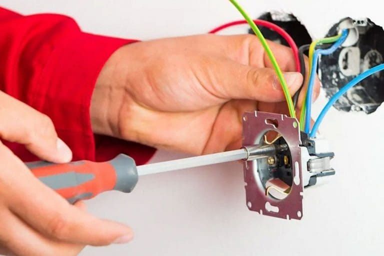 Una persona con una manga roja utiliza un destornillador para instalar o reparar una toma de corriente de pared con el cableado expuesto.