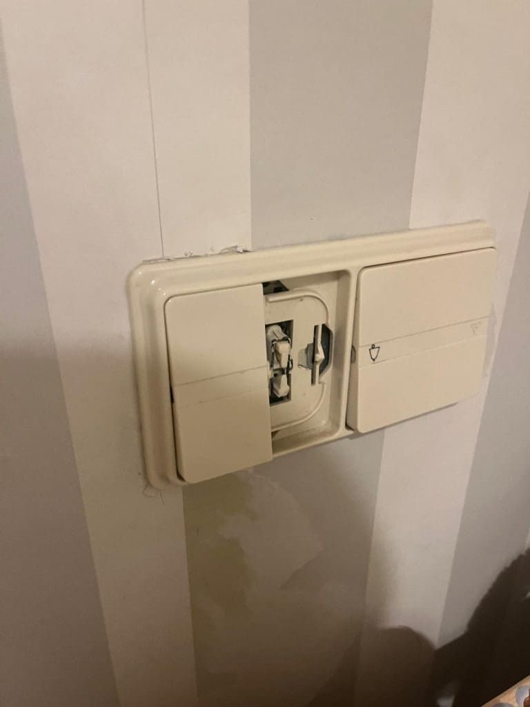 Una placa de interruptor de luz doble de color beige en una pared a rayas, a la que le falta una de las tapas de los dos interruptores basculantes.