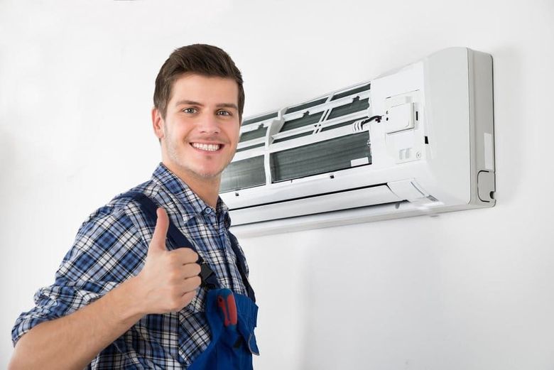 Un técnico sonriente, vestido con una camisa a cuadros y un mono azul, levanta el pulgar junto a un aire acondicionado montado en la pared.