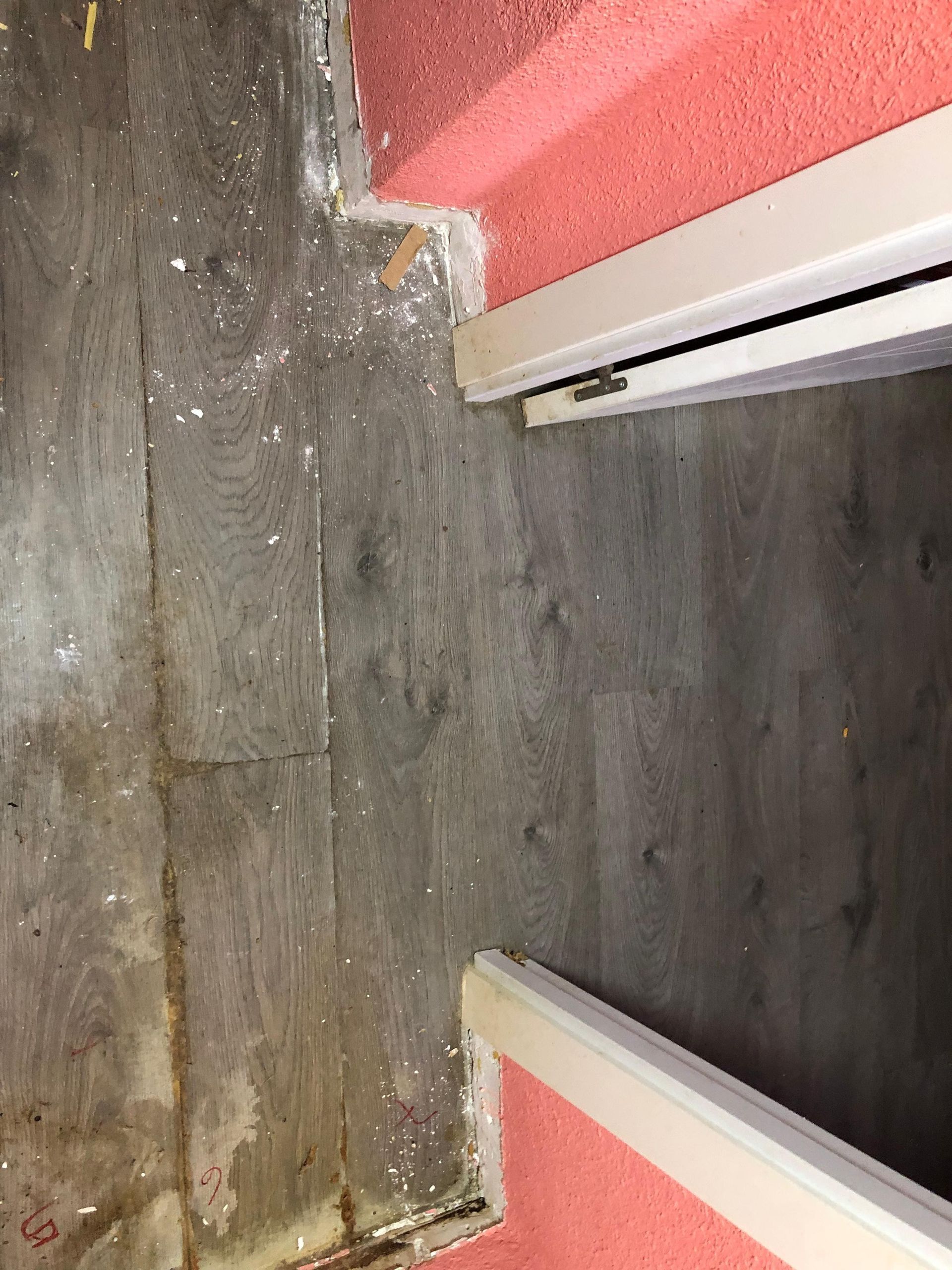 Una transición entre un suelo de madera desgastado y salpicado de pintura y un suelo limpio de color gris oscuro, divididos por el marco de una puerta.