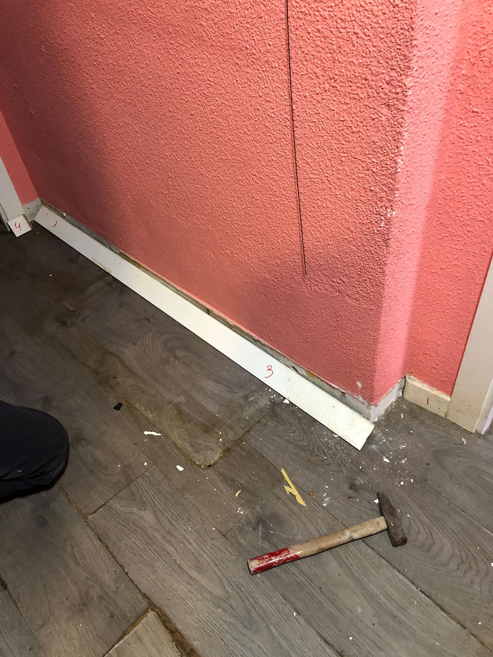Un martillo yace sobre un suelo de aspecto de madera oscura, junto a una pared rosa texturizada con una grieta vertical cerca del rodapié.