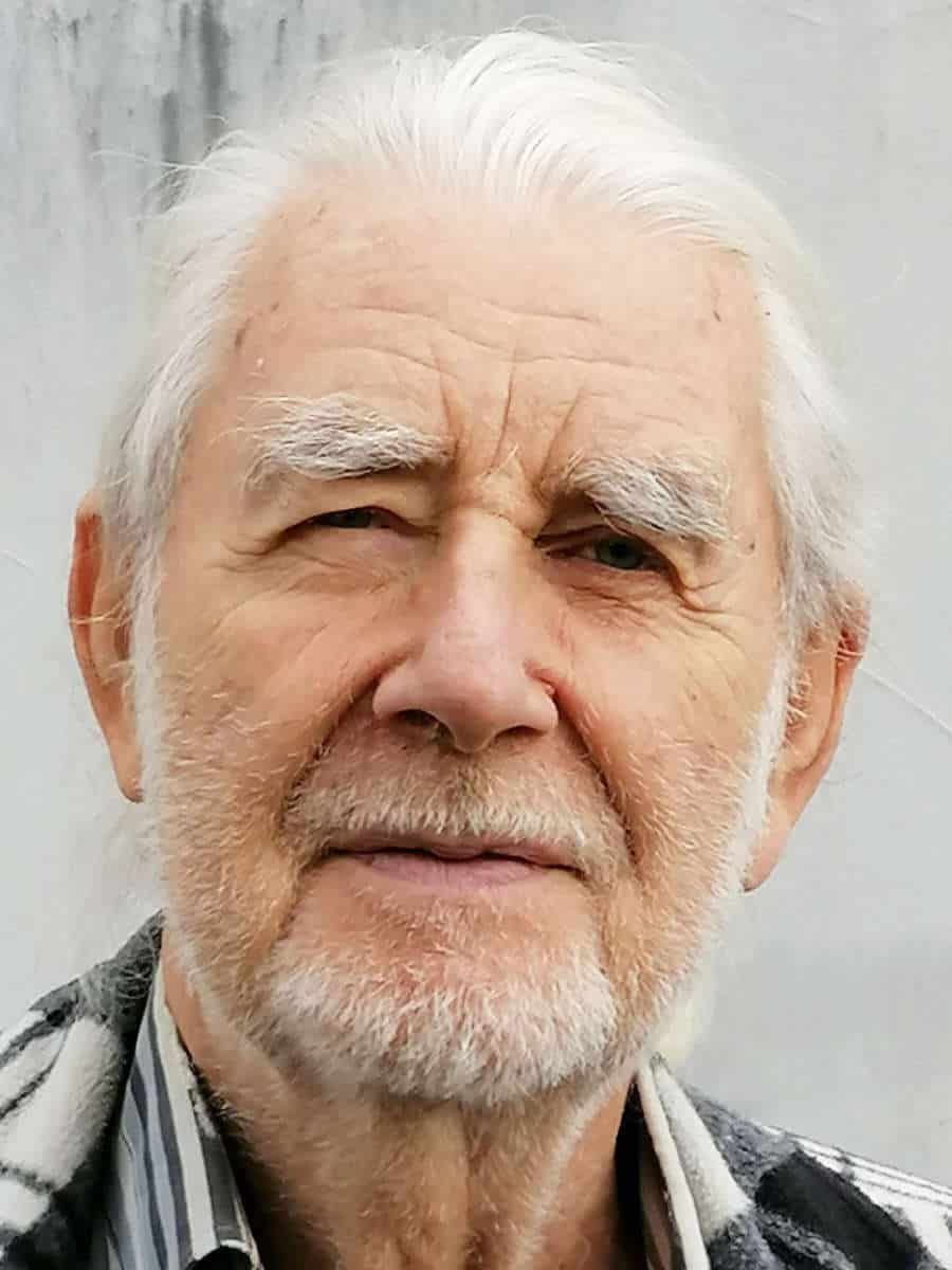 Ein älterer Mann mit grauem Haar und Bart schaut in die Kamera.