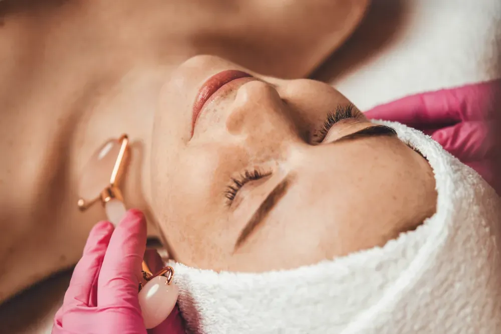 Una persona recibe un masaje facial con un rodillo de cuarzo rosa por parte de un profesional que usa guantes médicos rosas.