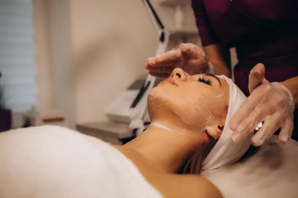 Una esteticista con guantes aplica un tratamiento facial a una clienta que lleva una diadema en un spa.