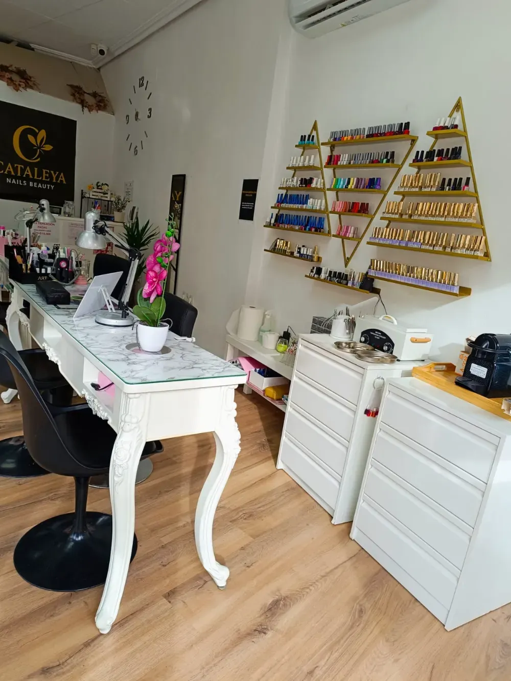 Un salón de uñas cuenta con un escritorio blanco, una silla con ruedas negra y estantes triangulares con marcos dorados