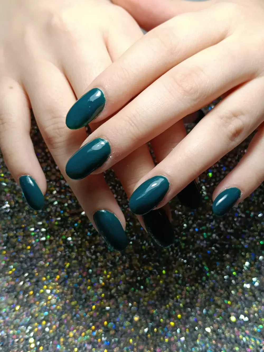 Manos con uñas de forma ovalada pintadas de un verde azulado oscuro brillante, apoyadas sobre una 