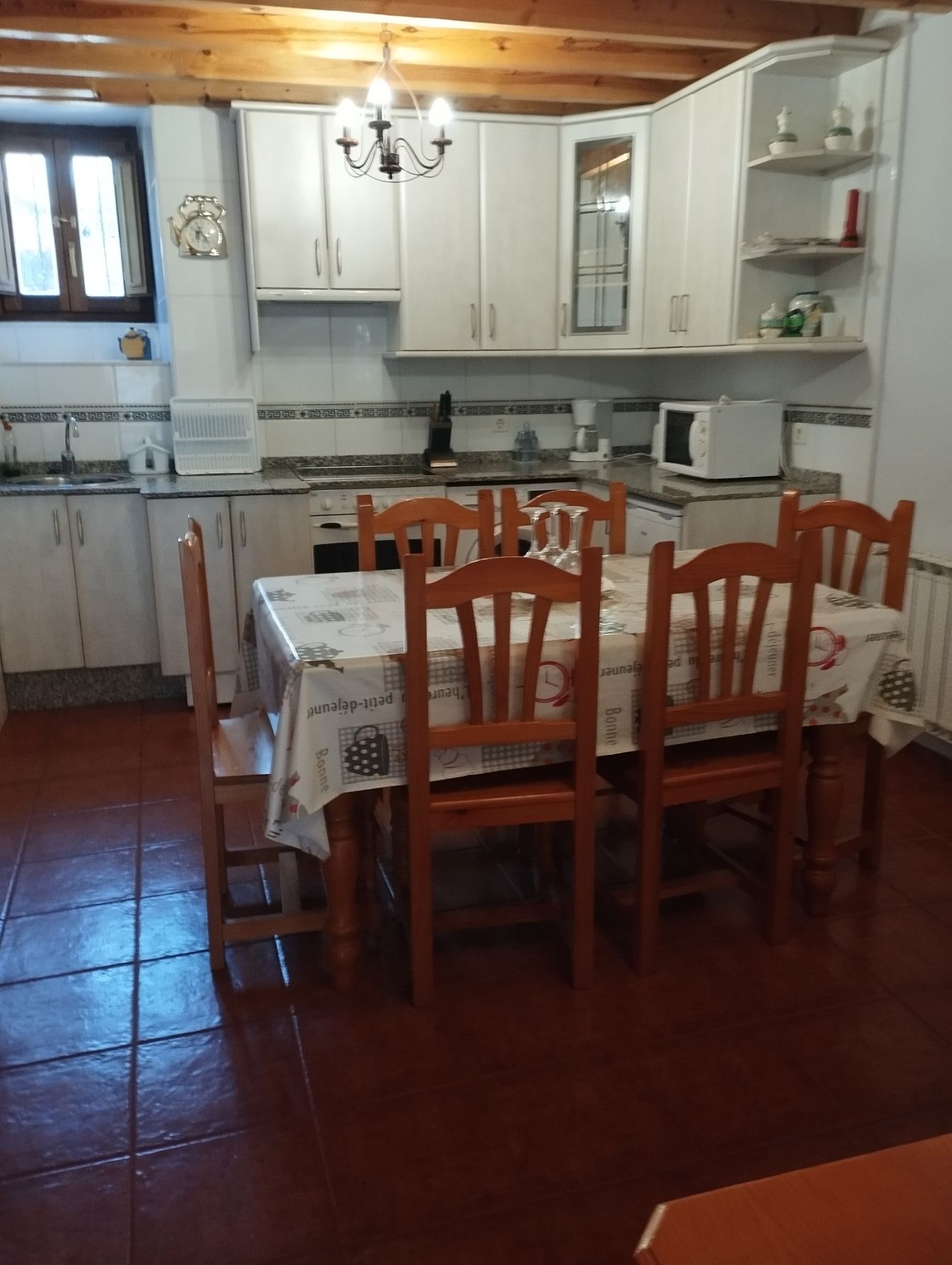 Cocina con armarios blancos, mesa y sillas de madera y suelo de baldosas rojas.