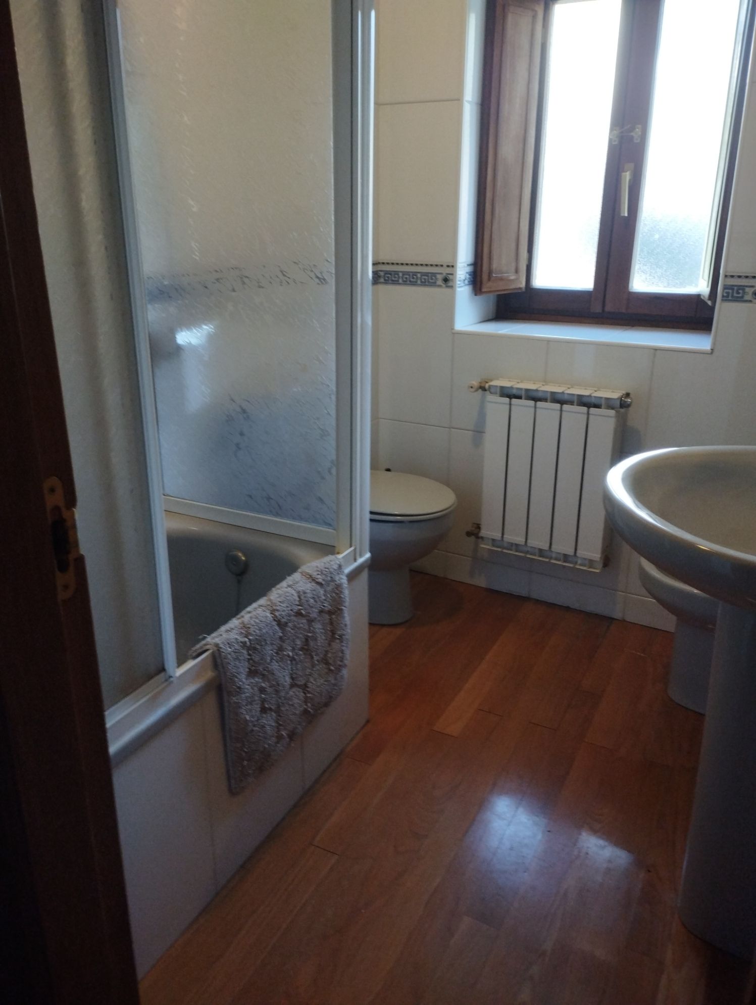Baño con bañera, inodoro, lavabo y radiador. Suelo de madera, ventana con persianas.