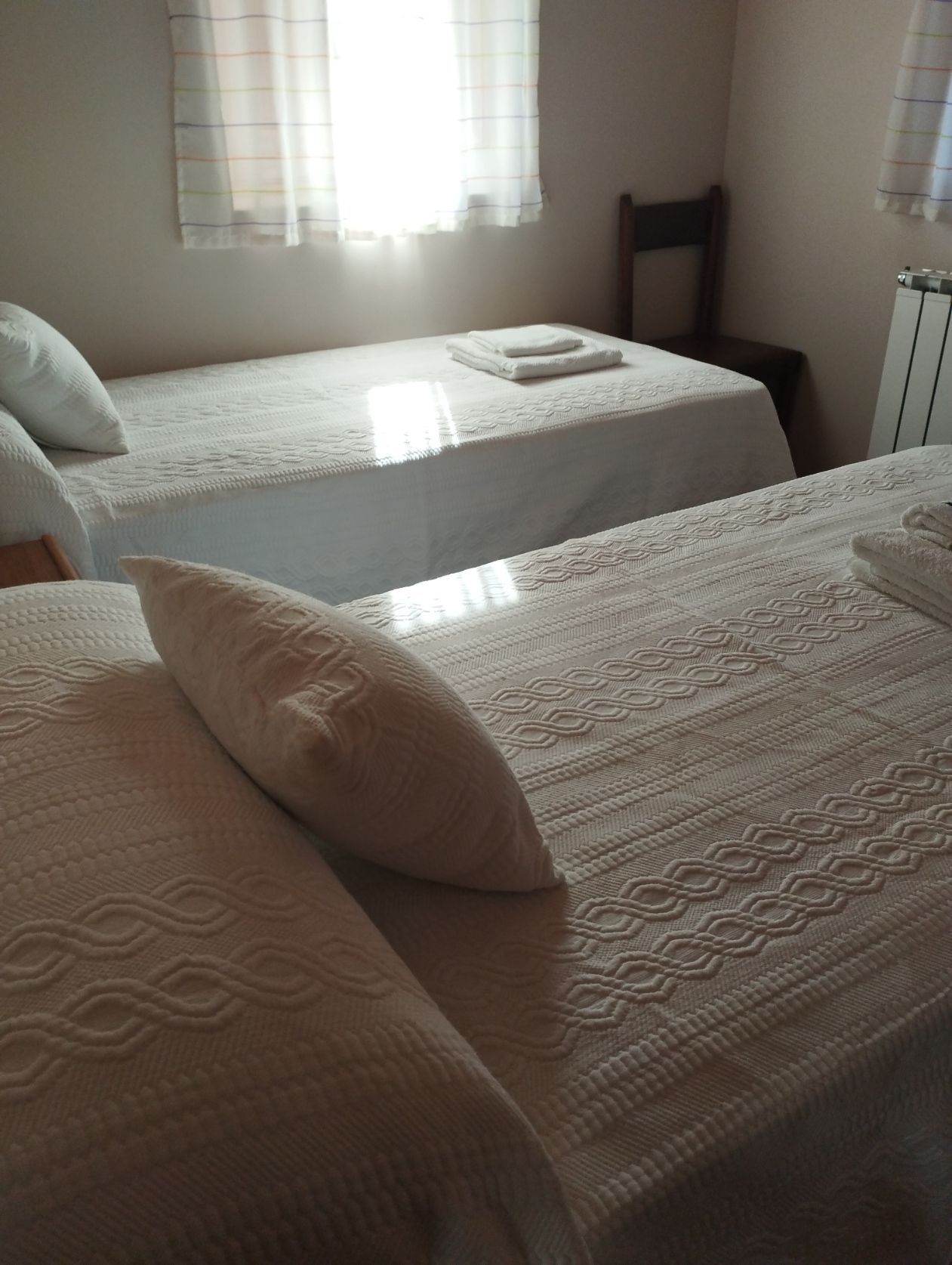 Dos camas individuales con sábanas y almohadas blancas en un dormitorio, con una ventana y una silla a la vista.