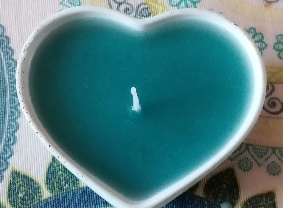Vela turquesa en forma de corazón en un portavelas blanco sobre un paño estampado.