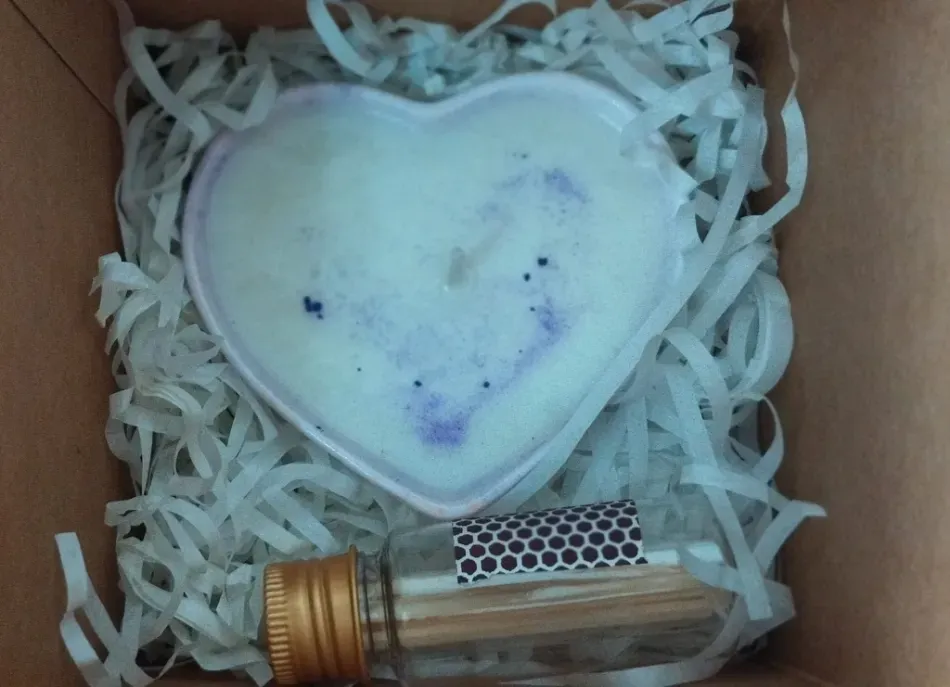 Jabón blanco en forma de corazón con motas azules en una caja de regalo sobre papel triturado, junto a un pequeño frasco de vidrio.