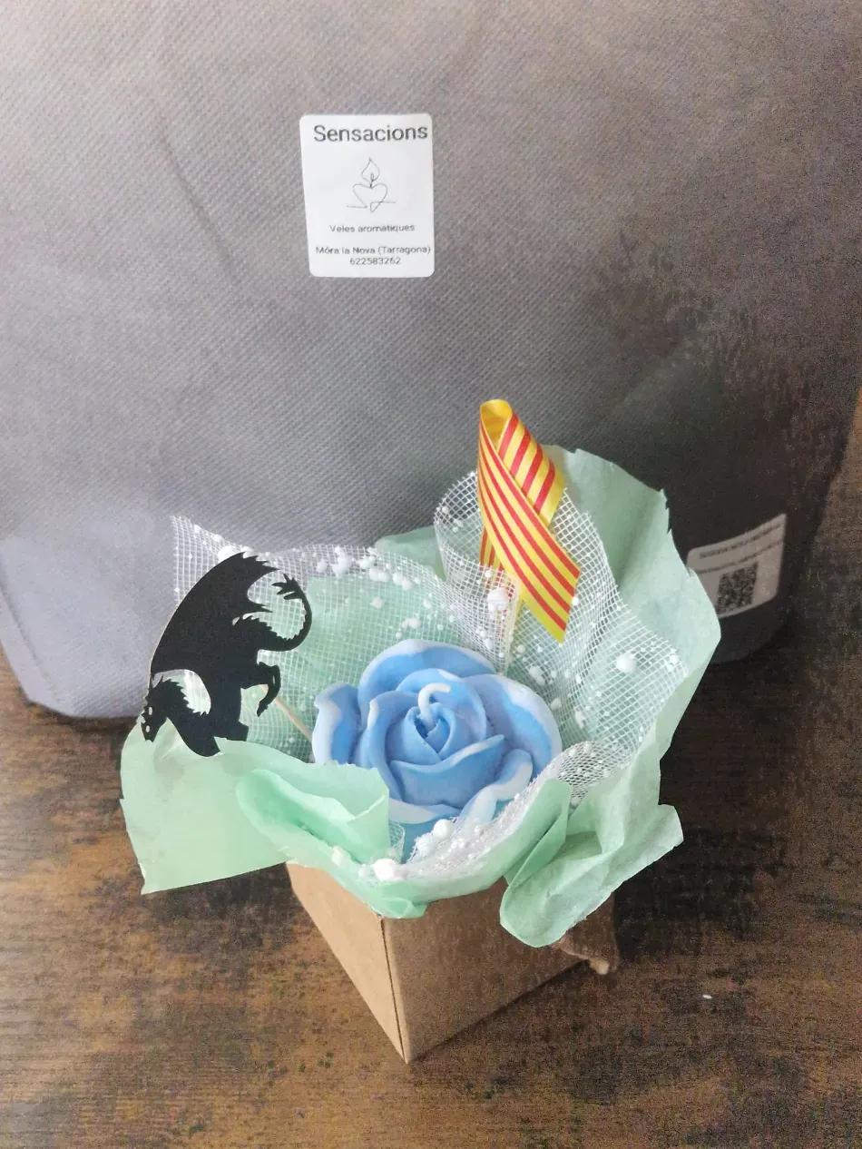 Pequeña caja de regalo con una rosa azul, papel verde, silueta negra y vela a rayas sobre una silla gris.