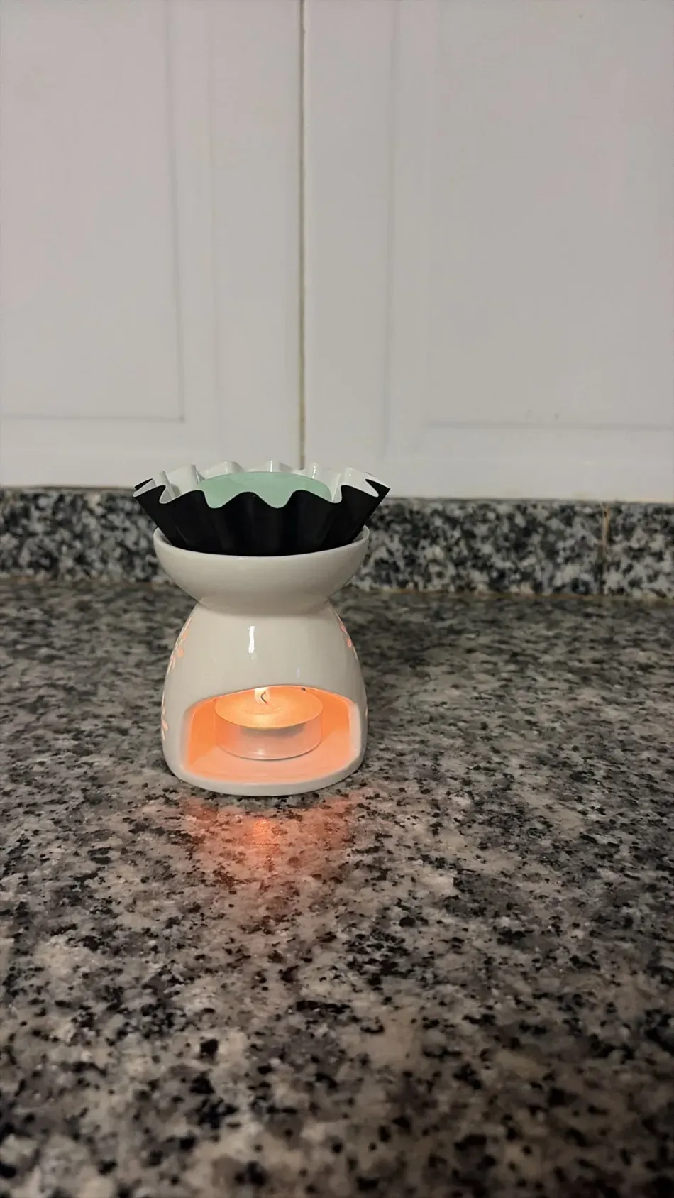 Un pequeño calentador de cera blanco sobre una encimera de granito moteado, que emite un brillo anaranjado, con un recipiente negro encima.