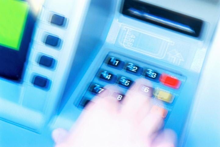 Mão de uma pessoa borrada ao digitar o PIN no teclado de um caixa eletrônico. Tons de azul com números de 1 a 9 visíveis.