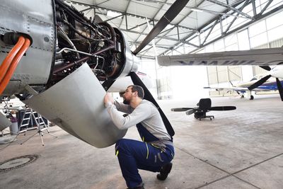 Un technicien en combinaison bleue est accroupi, effectuant la maintenance d'un moteur d'avion ouvert à l'intérieur d'un hangar.