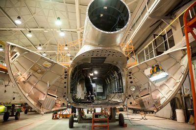 Dans un hangar industriel lumineux, le fuselage d'un gros avion est ouvert pour maintenance, et l'on aperçoit des ouvriers à l'intérieur.