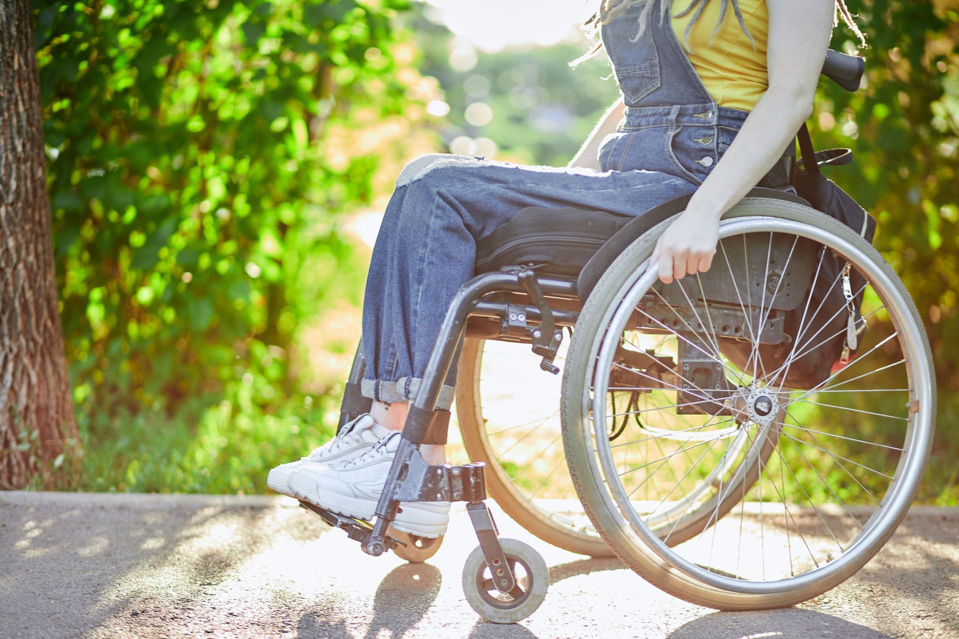 Mujer con overol de mezclilla en silla de ruedas en un camino soleado, con árboles verdes borrosos en el fondo.