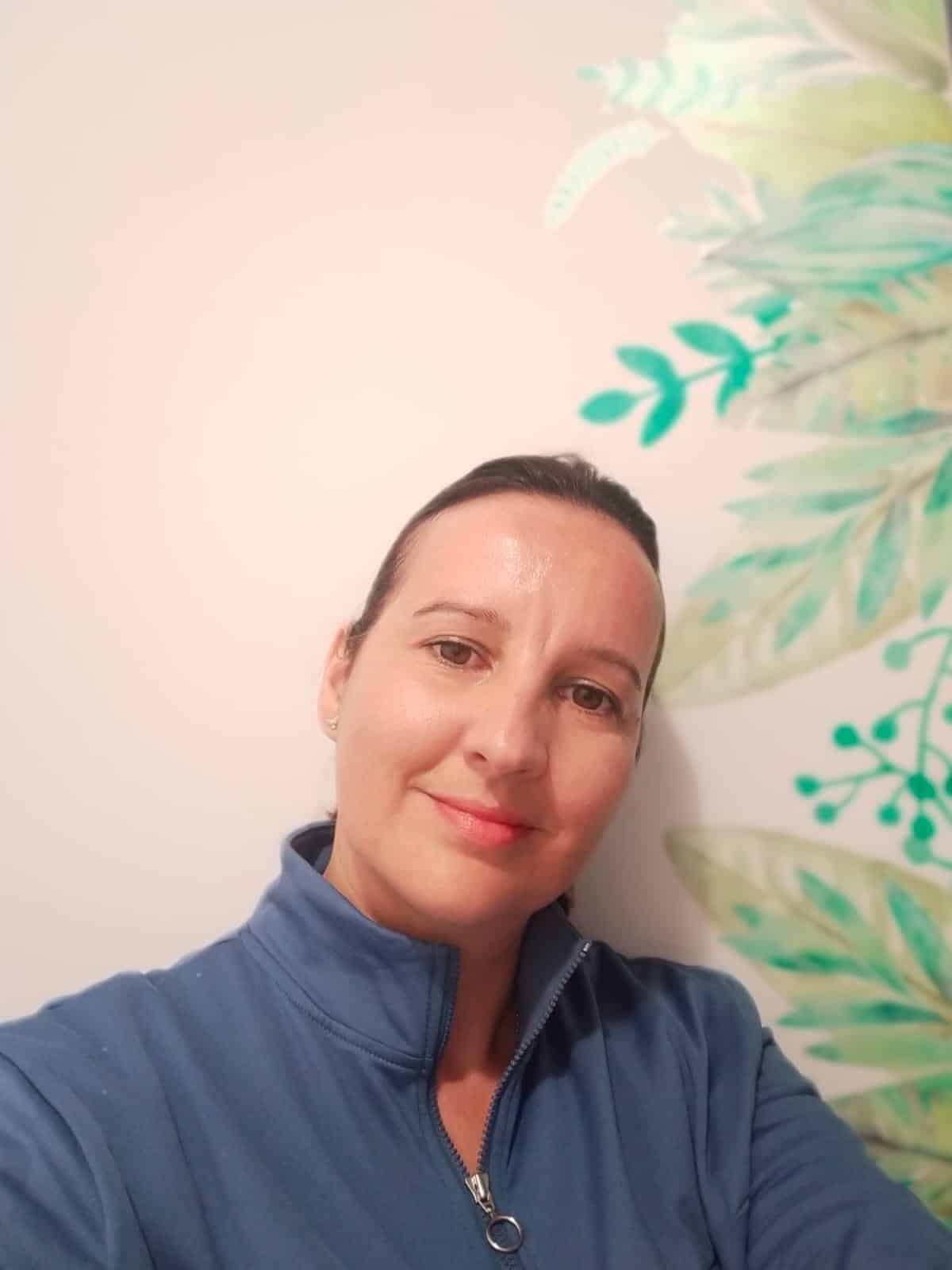 Una mujer de cabello castaño sonríe a la cámara, vistiendo una blusa azul, contra una pared con arte floral verde y blanco.