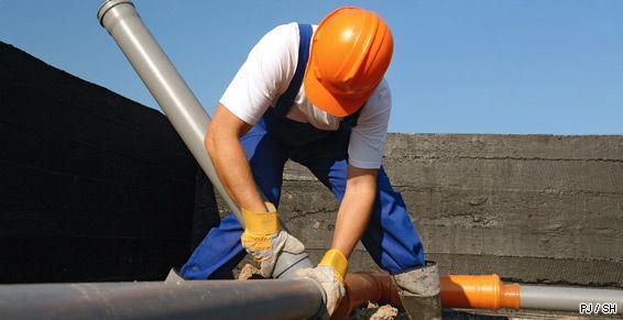 Ouvrier du bâtiment portant un casque orange soude un gros tuyau métallique à l'extérieur.