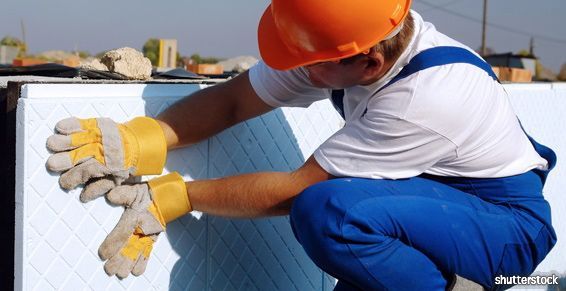 Un ouvrier du bâtiment, portant un casque orange et un pantalon bleu, est accroupi à côté d'une surface blanche isolée, utilisant des gants.