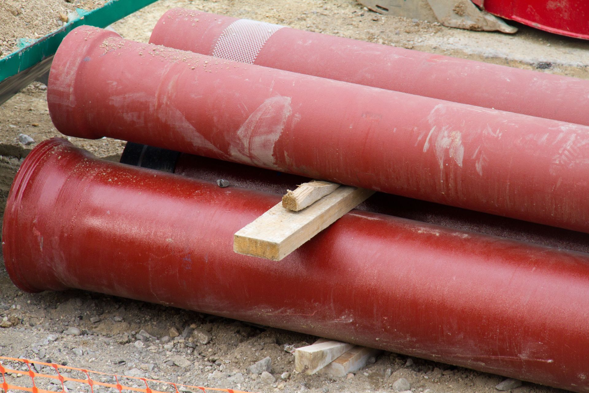 Des tuyaux métalliques rouges et roses empilés sur du gravier avec une cale en bois en dessous