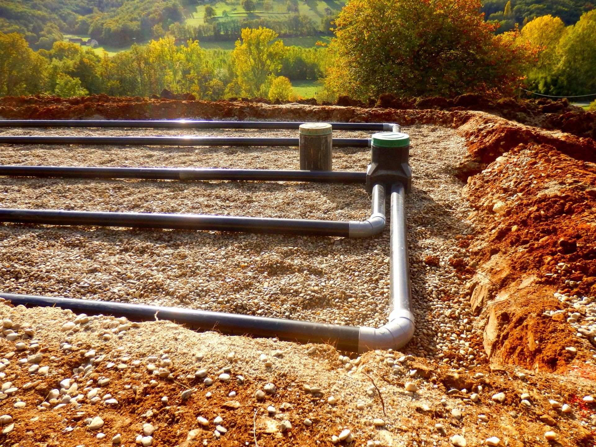 Des tuyaux d'irrigation traversent un champ de gravier, avec des arbres et des collines en arrière-plan.