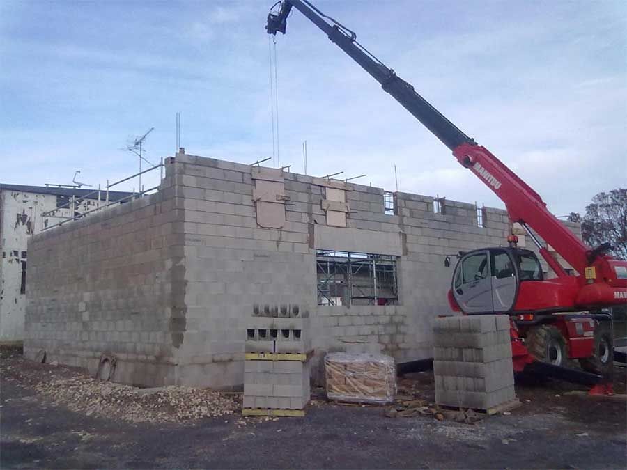 Une grue rouge soulève des blocs de béton sur un chantier de construction, à côté d'un immeuble en parpaings inachevé.
