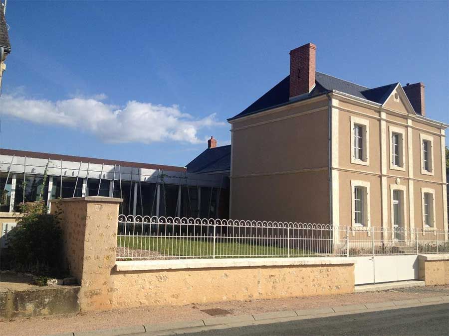 Bâtiment historique couleur sable avec des fenêtres à encadrement blanc, jouxtant une cour clôturée sous un ciel bleu.