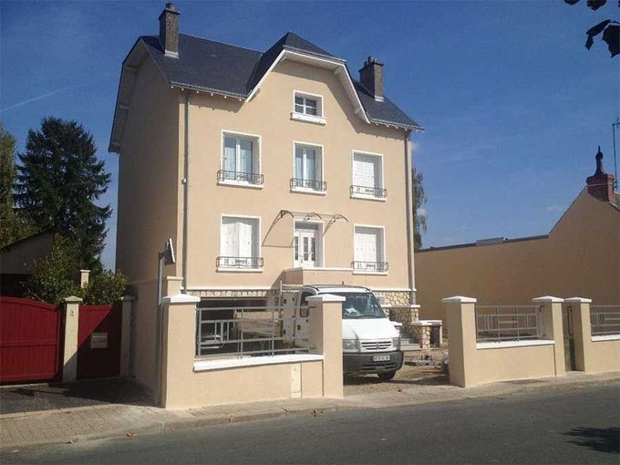 Maison beige de trois étages avec voiture garée et portail rouge dans une rue ensoleillée