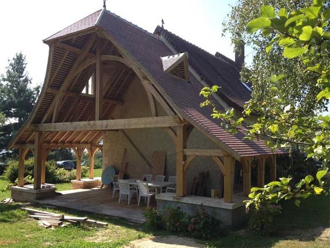Pavillon en bois avec un toit pentu en bardeaux et une terrasse ouverte, situé dans un jardin gazonné et arboré.