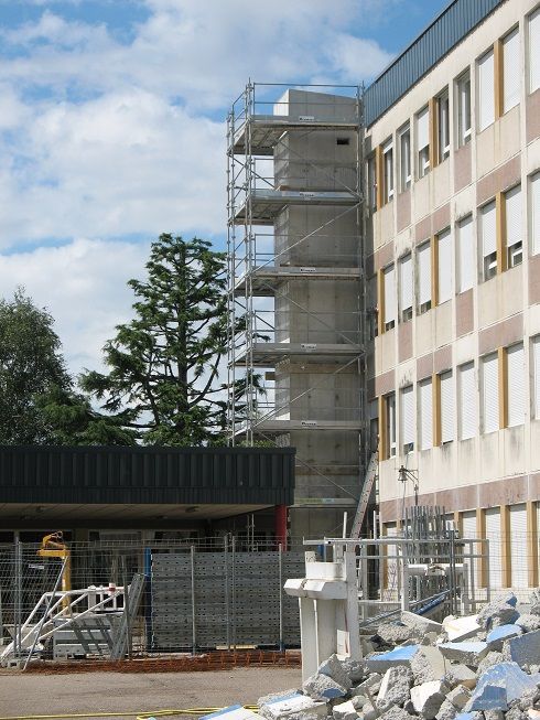 Échafaudages à côté d'un bâtiment beige, avec des débris et du matériel de construction au premier plan sous un ciel partiellement nuageux.