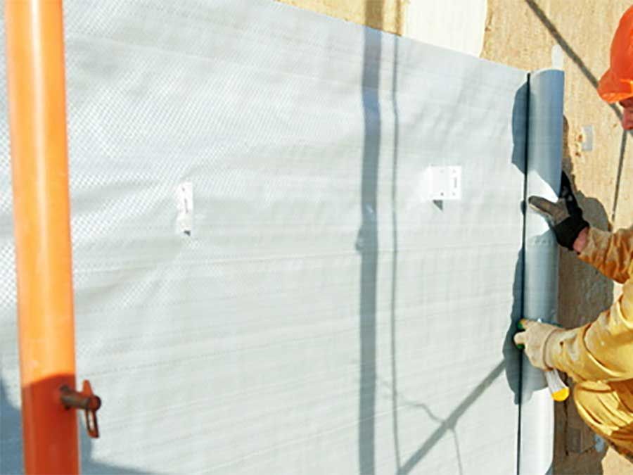 Des ouvriers fixent une bâche blanche à un mur, à côté de poteaux orange, à l'aide d'outils.