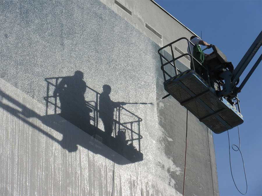 Des ouvriers dans une cabine d'ascenseur arrosent le mur d'un bâtiment, créant une tache humide et lumineuse ainsi que des ombres.