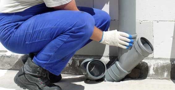Un ouvrier accroupi près de tuyaux en PVC gris ajuste un raccord bleu dans une zone technique.