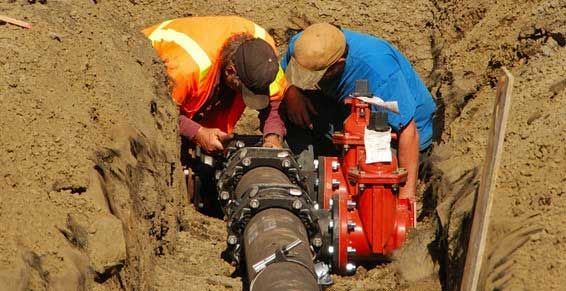 Deux ouvriers en vestes orange et bleues réparent une grosse canalisation dans une tranchée de terre.