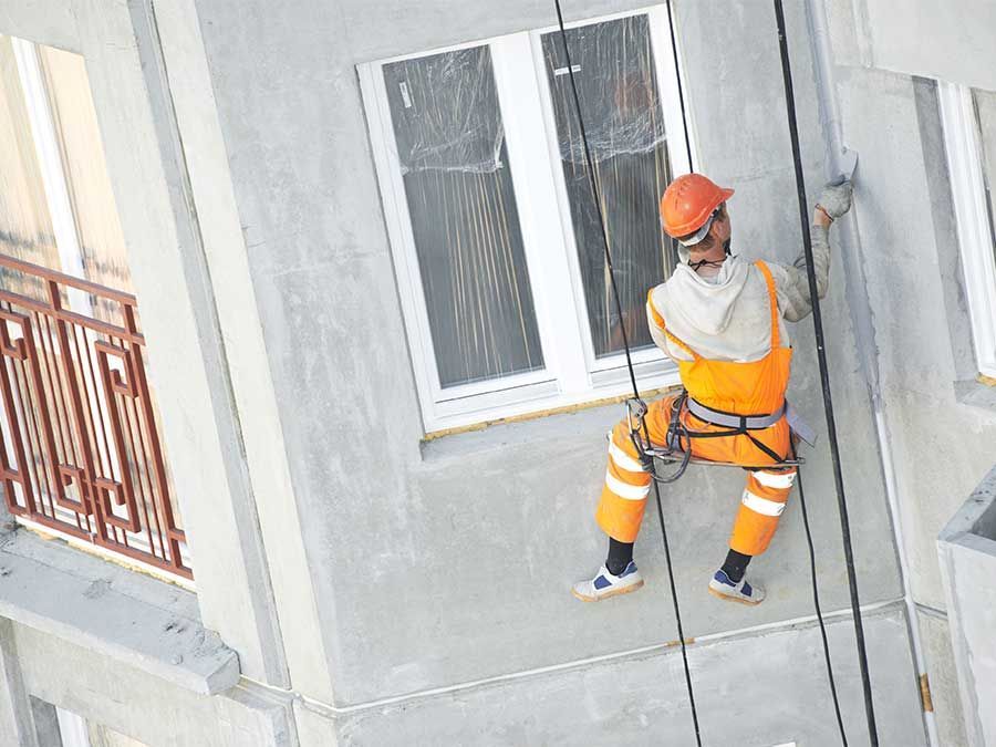 Un ouvrier en tenue de sécurité orange et casque est suspendu à des cordes près d'une fenêtre d'immeuble pendant qu'il travaille sur le mur extérieur.