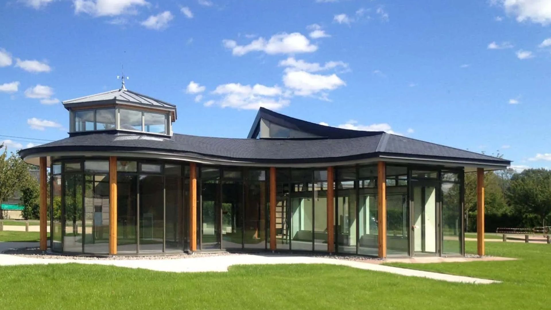 Pavillon moderne en verre avec un toit incurvé et une petite tour, situé sur une pelouse verte sous un ciel bleu.