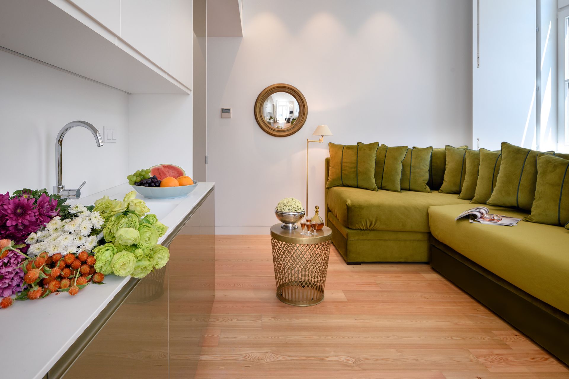 Small living space with olive green sofa, white countertops with flowers, and a round mirror.