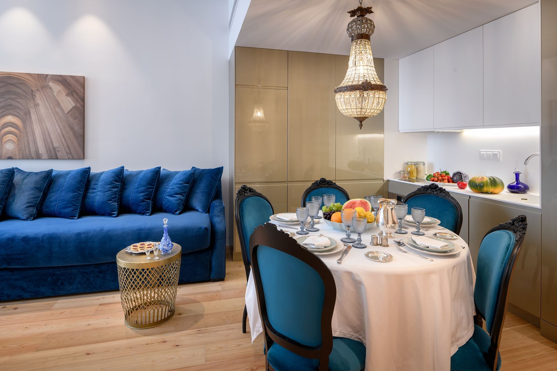 Blue sofa and table with teal chairs in a well-lit dining area, kitchen with fruit, gold accents, chandelier.