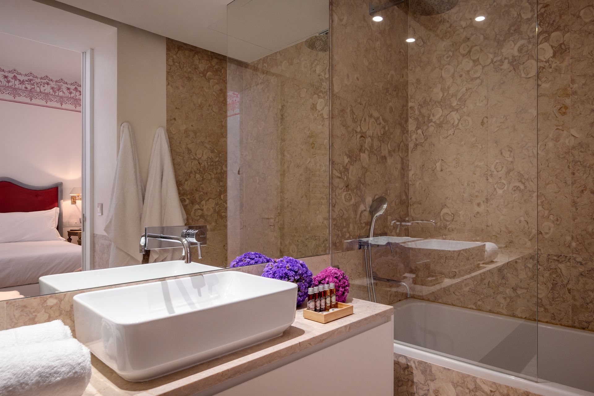 Bathroom with marble walls, white sink, and bathtub. The bedroom is visible in the background.