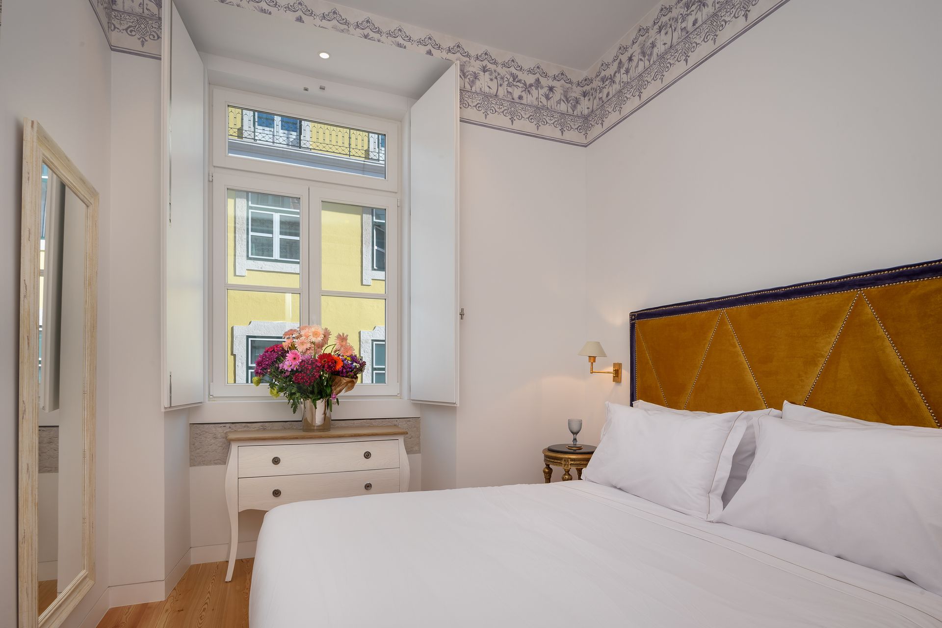 Bedroom with bed, yellow headboard, flowers in vase, and window.