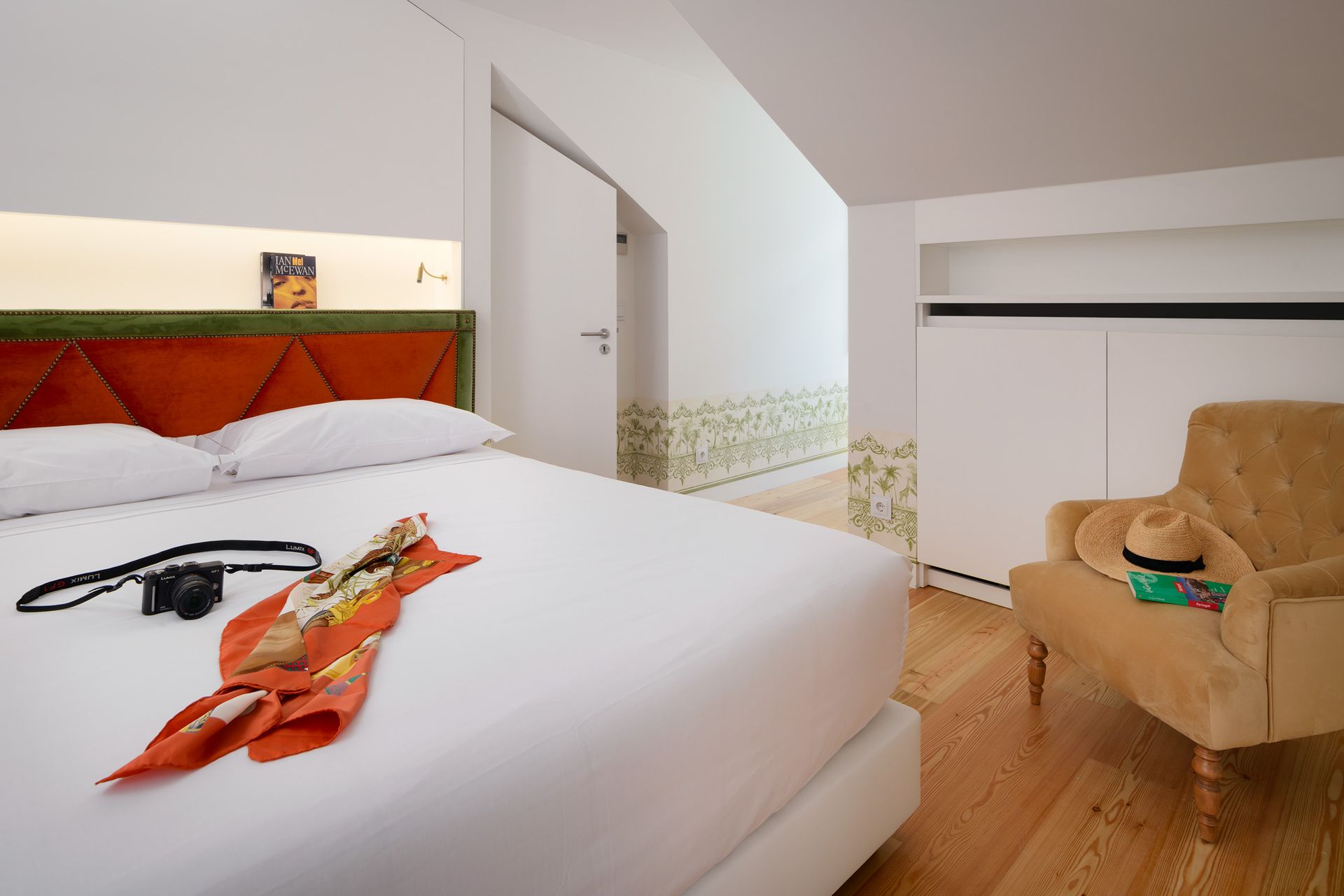 Bedroom with orange and green headboard, white bedding, armchair, wooden floor, and a scarf.