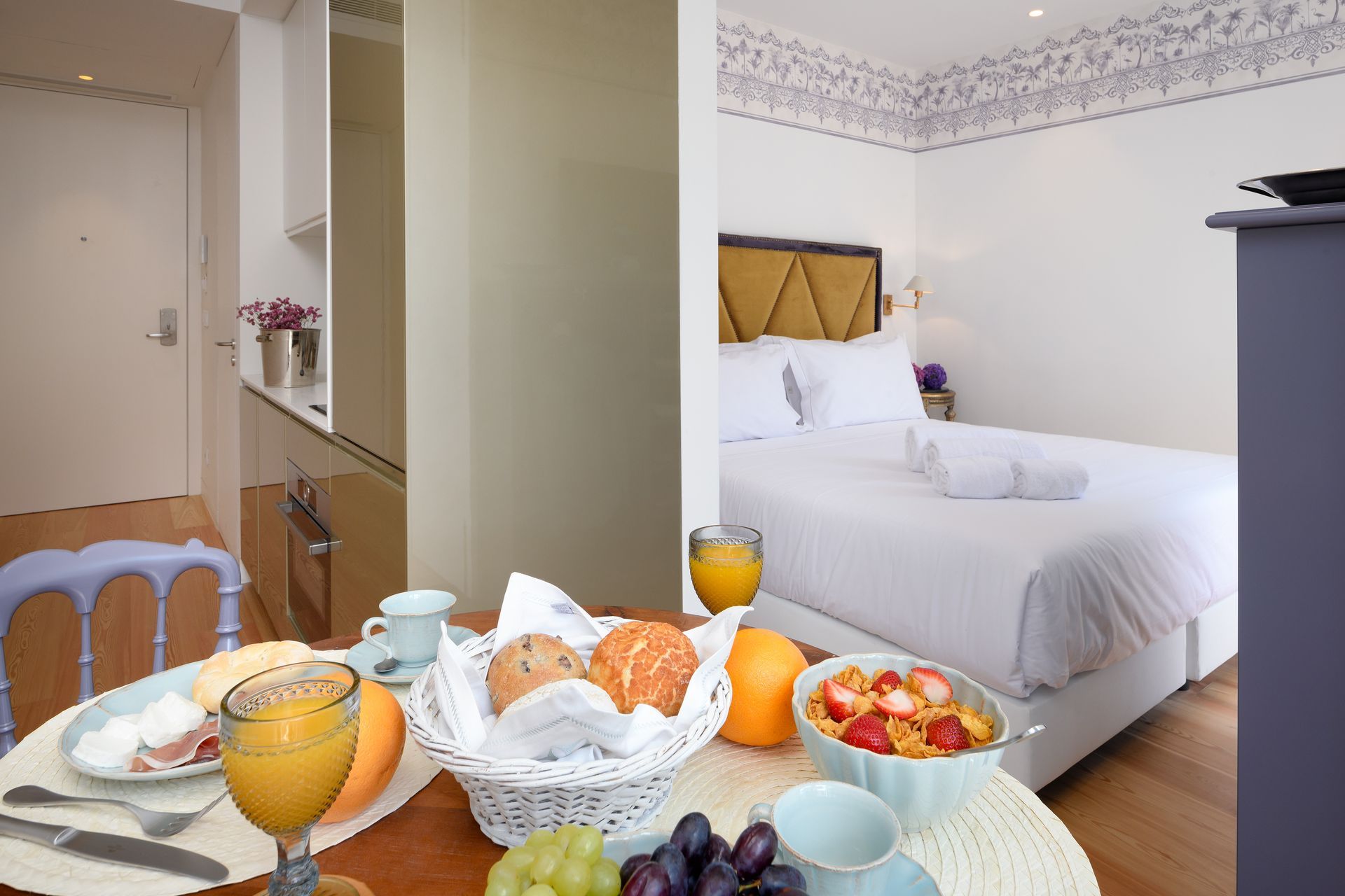 Breakfast table set in front of a small apartment bedroom with food, juice, and a bed with white linens.