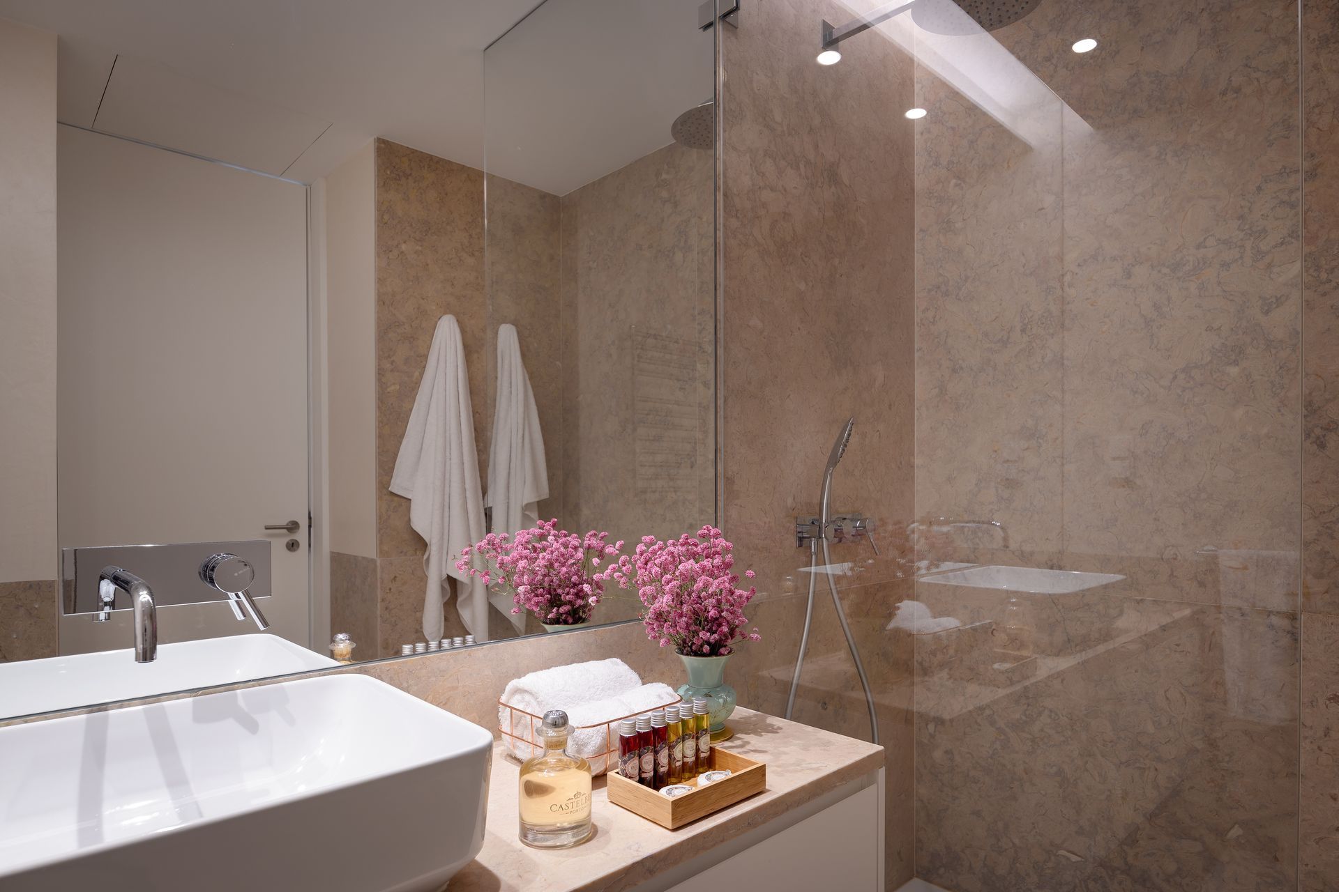 Modern bathroom with beige marble walls, glass shower, and white towels.