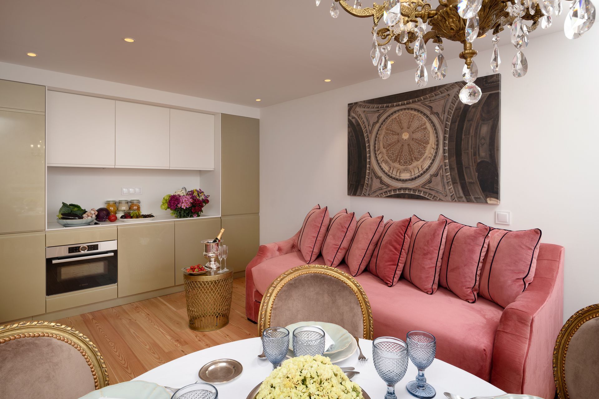 Pink sofa, dining table, and small kitchen in a well-lit space, with a mosaic art piece and chandelier.