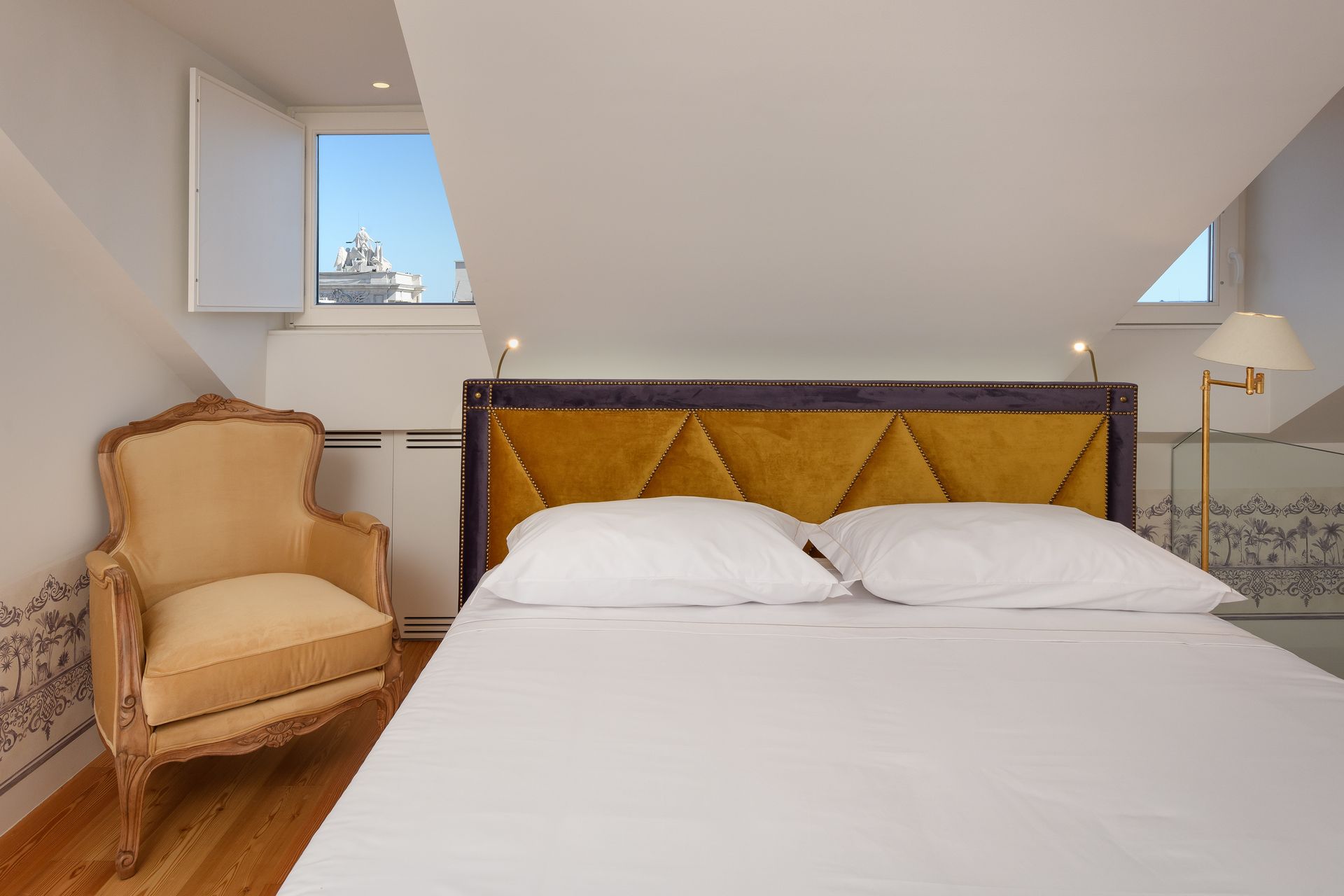 Bedroom with bed, yellow headboard, antique chair, and a view of buildings through a window.