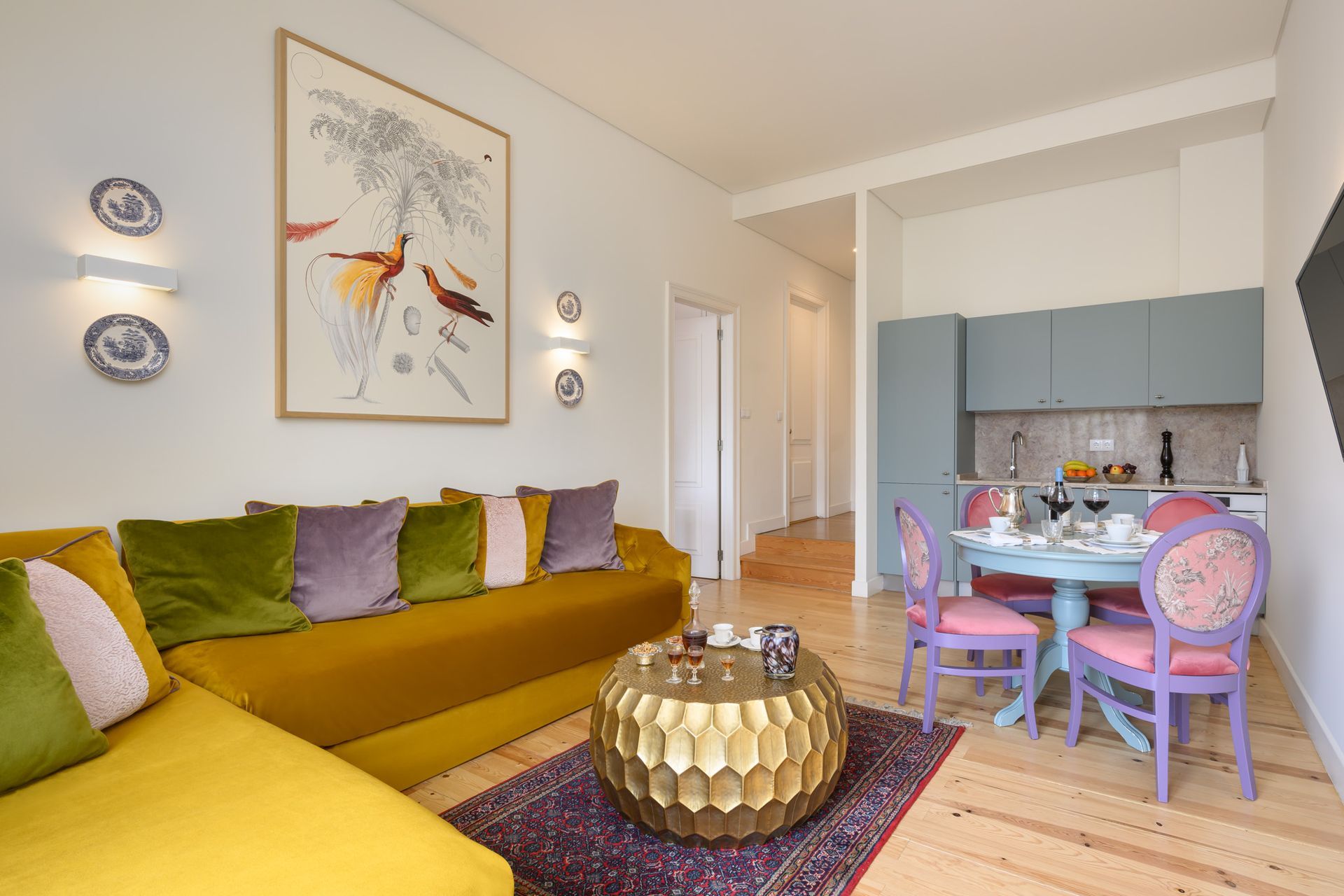 Living room with yellow sofa, purple chairs, and a round table. Artwork on the wall.