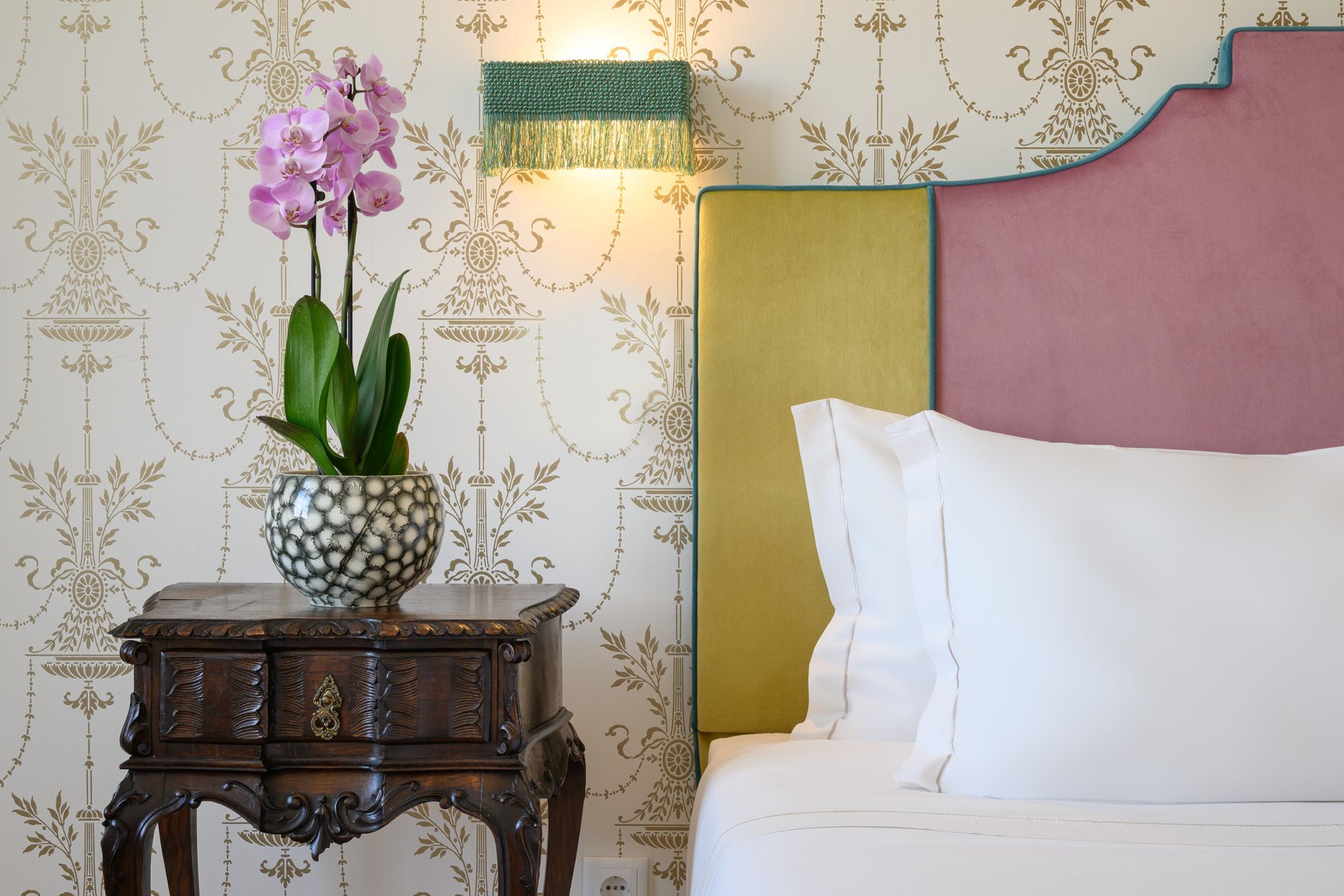 Bedroom with ornate headboard, nightstand with orchid, and patterned wallpaper.